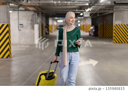 A woman wearing a hijab walks through a parking garage while pulling a suitcase and looking at her phone A woman wearing a hijab walks through a parking garage while pulling a suitcase and looking at her phone 135980815