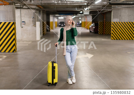 A smiling woman in a hijab is talking on her phone while standing in a parking garage with her yellow suitcase A smiling woman in a hijab is talking on her phone while standing in a parking garage with her yellow suitcase 135980832