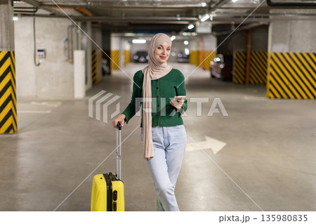 A woman wearing a hijab stands in a parking garage with her luggage, holding a phone A woman wearing a hijab stands in a parking garage with her luggage, holding a phone 135980835