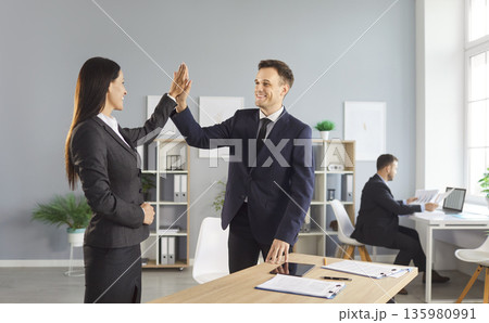 Two smiling business colleagues standing in office, giving each other high five, celebrating success Two smiling business colleagues standing in office, giving each other high five, celebrating success 135980991