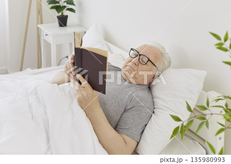 Gray-haired senior elderly man in eyeglasses reading a book lying on bed in his bedroom at home Gray-haired senior elderly man in eyeglasses reading a book lying on bed in his bedroom at home 135980993