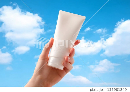 Hand of woman is holding a white plastic tube container for moisturizer or sunscreen on blue sky. Cosmetic bottle for design packaging. Minimal scene for advertising Hand of woman is holding a white plastic tube container for moisturizer or sunscreen on blue sky. Cosmetic bottle for design packaging. Minimal scene for advertising 135981249