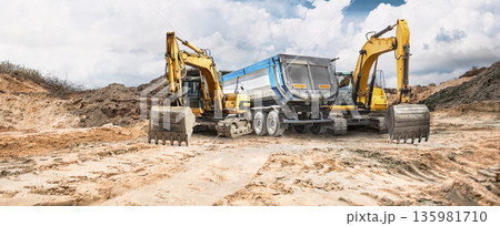 Construction activity is ongoing on dry, cracked earth where excavators are seen positioned beside a truck under a partly cloudy sky Construction activity is ongoing on dry, cracked earth where excavators are seen positioned beside a truck under a partly cloudy sky 135981710