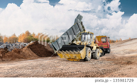 A loader lifts and unloads material at a construction site next to a dump truck. The sky is overcast. Construction work is underway below 135981729
