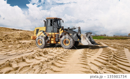 Deep tracks from heavy machinery cut through sandy ground as a loader works near a large sand pile under a cloudy sky during construction activity Deep tracks from heavy machinery cut through sandy ground as a loader works near a large sand pile under a cloudy sky during construction activity 135981734