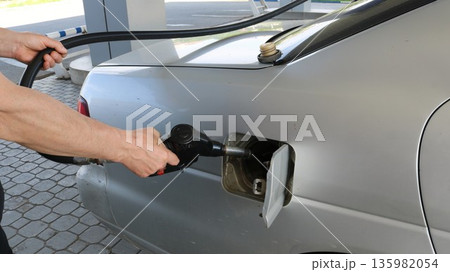 A white man's hands hold a dispensing nozzle near the open gas tank of a gray car while refueling it at a petrol station. Filling the tank with high-quality gasoline at a Russian gas station. A white man's hands hold a dispensing nozzle near the open gas tank of a gray car while refueling it at a petrol station. Filling the tank with high-quality gasoline at a Russian gas station. 135982054