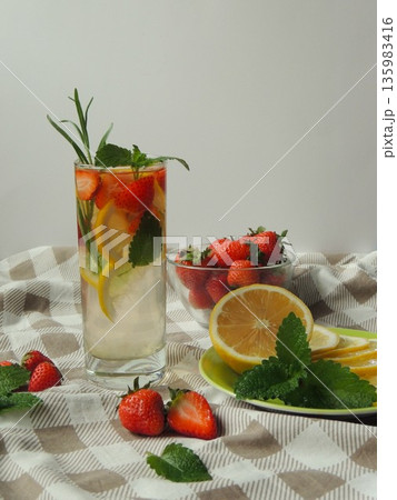 A tall glass of refreshing citrus-strawberry-mint lemonade and a natbrmort of fresh berries, lemon and herb leaves on a checkered light tablecloth on a gray background 135983416