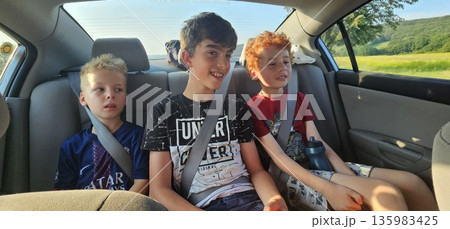 Three young boys sit buckled in the back seat of a car during a sunny countryside drive, smiling and chatting Three young boys sit buckled in the back seat of a car during a sunny countryside drive, smiling and chatting 135983425