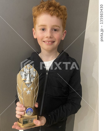 Smiling young boy holding a trophy indoors, celebrating a win or achievement. 135983430
