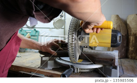 Cutting a wooden piece on a miter saw, view from behind a carpenter, working on an end table with wood, an end cut on a wood product made on a machine in a workshop 135985506