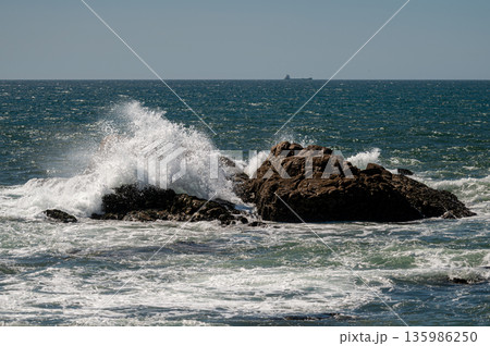 Ocean waves splashing against rocky formations while a cargo ship sails in the distance. 135986250