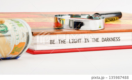 A magnifying glass rests on an open book displaying the message BE THE LIGHT IN THE DARKNESS 135987488