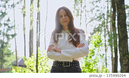 Confident young woman with crossed arms stands in sunlight outdoors, expressing empowerment, strength, and modern feminine style. Confident young woman with crossed arms stands in sunlight outdoors, expressing empowerment, strength, and modern feminine style. 135990204