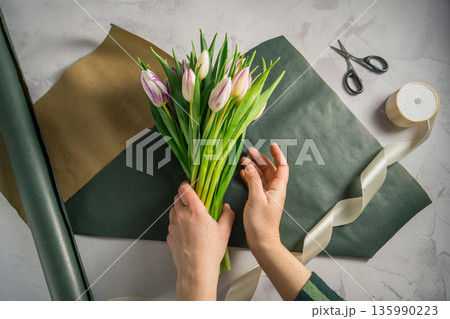 Mother's day, birthday bouquet. Female hands wrapping tulip flowers in craft paper. Spring tulips flower background 135990223