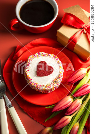 Valentine's Day festive red background. Cake with red heart, cup of coffee, gift box and spring red tulip flower bouquet. Top view Valentine's Day festive red background. Cake with red heart, cup of coffee, gift box and spring red tulip flower bouquet. Top view 135990294