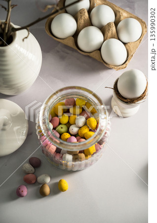 Easter background with mini egg candies in a jar, twigs in a vase and white chicken eggs Easter background with mini egg candies in a jar, twigs in a vase and white chicken eggs 135990302