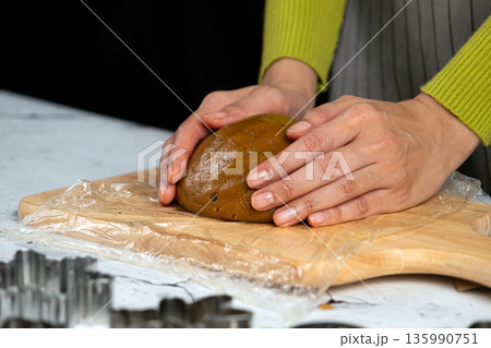 Close-up of hands kneading cookie dough on a wooden cutting board with plastic wrap, preparing homemade baked goods in a cozy kitchen 135990751