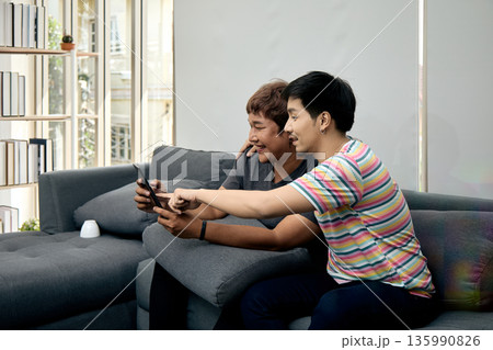 Two Asian men sitting on sofa smiling and pointing at tablet screen in cozy home. This candid moment reflects digital connection, friendship, and modern tech lifestyle 135990826