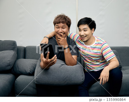 Two Asian LGBTQ men sitting close on sofa, smiling and enjoying content on tablet together. This cheerful indoor scene highlights friendship, connection, and digital lifestyle Two Asian LGBTQ men sitting close on sofa, smiling and enjoying content on tablet together. This cheerful indoor scene highlights friendship, connection, and digital lifestyle 135990864