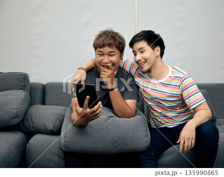 Two LGBTQ Asian men sitting on sofa, smiling and enjoying watching content on tablet together. This moment reflects digital bonding, friendship, and relaxed lifestyle at home Two LGBTQ Asian men sitting on sofa, smiling and enjoying watching content on tablet together. This moment reflects digital bonding, friendship, and relaxed lifestyle at home 135990865