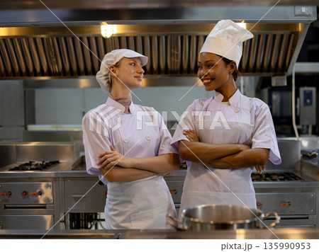 Two women chef coworkers standing confidently with crossed arms in professional kitchen smiling at each other; teamwork leadership hospitality career pride Two women chef coworkers standing confidently with crossed arms in professional kitchen smiling at each other; teamwork leadership hospitality career pride 135990953
