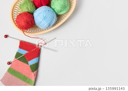 Red, green, blue and pink knitting balls in basket with knitting needles and knitted scarf on gray background. Set for the hobby of knitting warm clothes or toys. Concept needlework 135991143