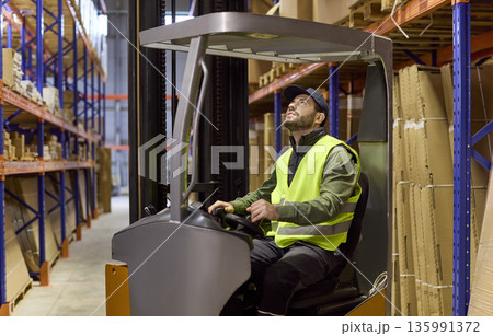 Warehouse worker driving forklift and checking shelves during pallet loading Warehouse worker driving forklift and checking shelves during pallet loading 135991372