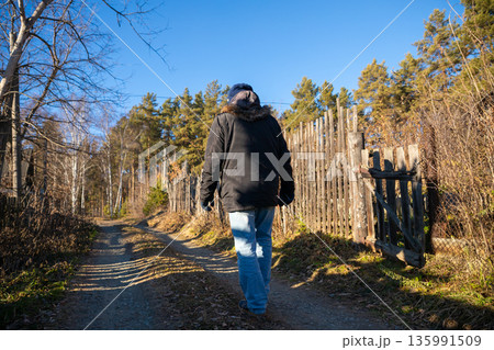 Man walks along a dirt road in a village with a wooden fence and trees on a sunny day. Rural nature and outdoor adventure. 135991509