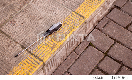Screwdriver with magnetic tip on a yellow tactile paving on a sidewalk. Tool for repair and construction. 135991662