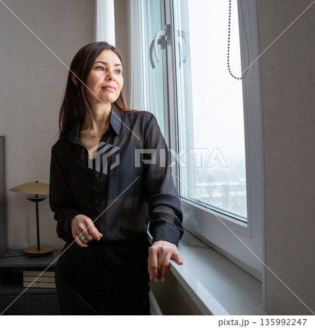 Woman stands thoughtfully by apartment window looking outside, a moment of reflection and calm for a successful businesswoman at home, cozy and modern room. realtor in her office Woman stands thoughtfully by apartment window looking outside, a moment of reflection and calm for a successful businesswoman at home, cozy and modern room. realtor in her office 135992247
