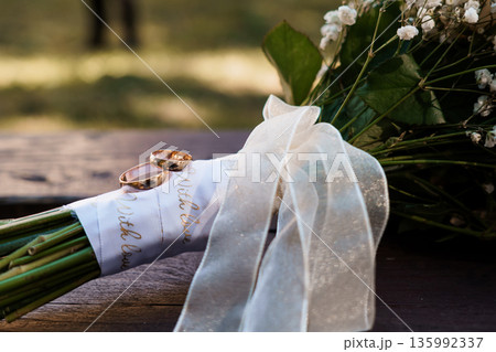 Golden wedding rings resting on white ribbon-wrapped bouquet with baby's breath flowers in soft natural light Golden wedding rings resting on white ribbon-wrapped bouquet with baby's breath flowers in soft natural light 135992337