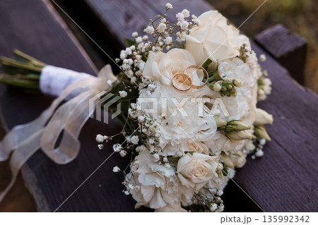 Elegant White Wedding Bouquet with Gold Rings on Wooden Bench in Soft Natural Light Elegant White Wedding Bouquet with Gold Rings on Wooden Bench in Soft Natural Light 135992342