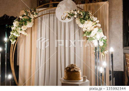 Elegant Wedding Arch Decorated with White Flowers and Greenery Under Soft Lighting with Cake Display 135992344