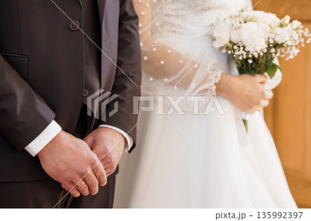 Close-up of bride and groom holding hands at wedding ceremony with white bouquet and elegant attire Close-up of bride and groom holding hands at wedding ceremony with white bouquet and elegant attire 135992397