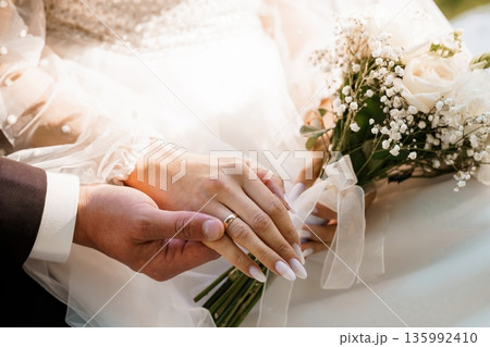 Close-up of bride and groom holding hands with wedding bouquet and gold rings on fingers, soft white lace dress, delicate flowers, romantic moment 135992410