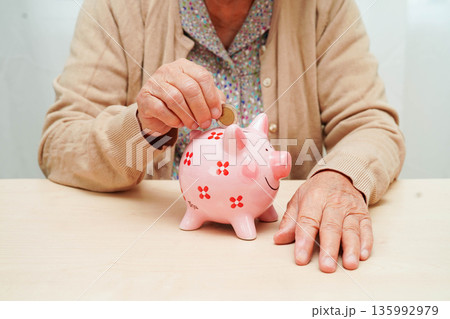 Retired elderly woman putting coins money in piggy bank and worry about monthly expenses and treatment fee payment. Retired elderly woman putting coins money in piggy bank and worry about monthly expenses and treatment fee payment. 135992979