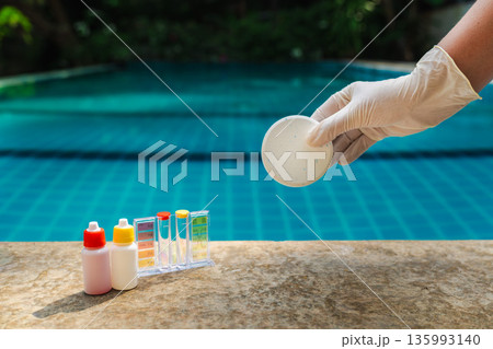 Gloved hand holding chlorine tablet above swimming pool with water testing kit and chemical bottles on stone ledge, tropical outdoor pool maintenance scene Gloved hand holding chlorine tablet above swimming pool with water testing kit and chemical bottles on stone ledge, tropical outdoor pool maintenance scene 135993140