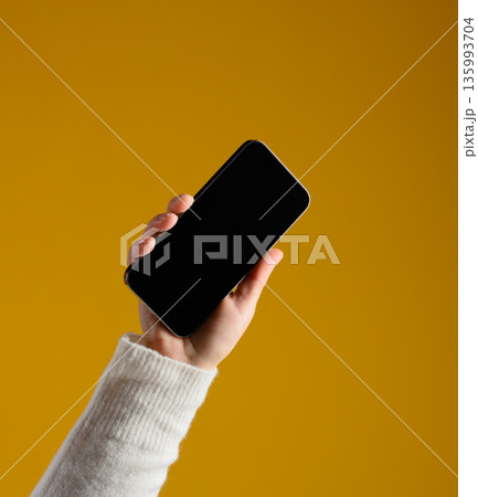 A man's hand in a white sweater holds a modern smartphone with a black screen on a rich yellow background. 135993704