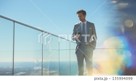 Confident young businessman in slim gray suit walking on rooftop terrace while using smartphone, surrounded by dreamy colorful bokeh and clear blue sky Confident young businessman in slim gray suit walking on rooftop terrace while using smartphone, surrounded by dreamy colorful bokeh and clear blue sky 135994099