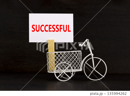 Successful symbol. Concept word Successful on beautiful white paper on clothespin. Beautiful black background. Beautiful miniature white bicycle. Business successful concept. Copy space. Successful symbol. Concept word Successful on beautiful white paper on clothespin. Beautiful black background. Beautiful miniature white bicycle. Business successful concept. Copy space. 135994262