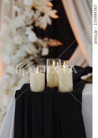 Three glass candles on black fabric table at wedding ceremony with floral background Three glass candles on black fabric table at wedding ceremony with floral background 135994367