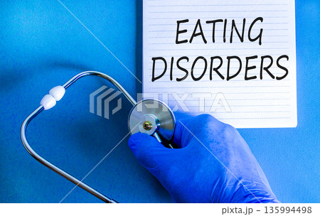 Eating disorders symbol. Concept words Eating disorders on beautiful white note. Doctor's hand in blue medical glove. Beautiful blue background. Medical healthy lifestyle eating disorders concept. Eating disorders symbol. Concept words Eating disorders on beautiful white note. Doctor's hand in blue medical glove. Beautiful blue background. Medical healthy lifestyle eating disorders concept. 135994498