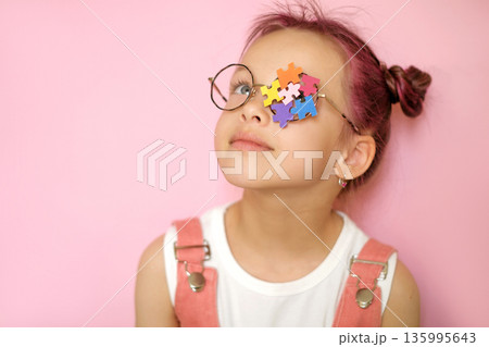 Young girl wearing glasses with colorful puzzle pieces covering one eye. Concept of autism awareness, neurodiversity, child development, education and inclusion on pink background. 135995643