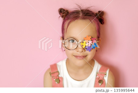 Portrait of a young girl wearing glasses with colorful puzzle pieces covering one eye. Visual concept of autism awareness, inclusion, neurodiversity, empathy and child development in studio setting. 135995644