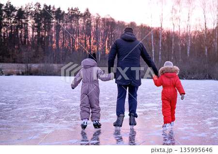 Rear view of father holding hands with two children while ice skating on frozen lake at sunset. Winter family leisure, parenting, outdoor bonding, childhood memories and seasonal lifestyle. 135995646