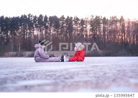 Two children resting on frozen lake after ice skating. Winter outdoor activity, childhood leisure, safety moment, playful pause during cold seasonal recreation in nature. 135995647