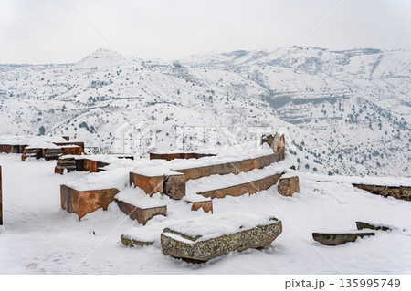 Snow-covered stone ruins stand near the Garni Temple, broken walls and ancient blocks contrast with the wide winter mountain landscape Snow-covered stone ruins stand near the Garni Temple, broken walls and ancient blocks contrast with the wide winter mountain landscape 135995749
