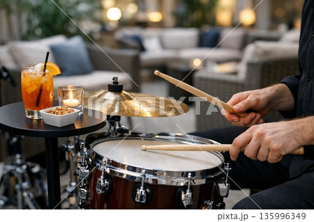 Man playing drums with drumsticks in a cozy indoor setting. Evening entertainment and live music concept for hospitality. Vibrant cocktail, candle Man playing drums with drumsticks in a cozy indoor setting. Evening entertainment and live music concept for hospitality. Vibrant cocktail, candle 135996349
