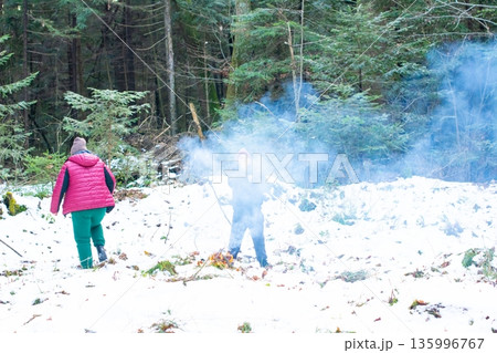 Two individuals hike through a snowy forest, smoke billowing from a small fire they've built. Two individuals hike through a snowy forest, smoke billowing from a small fire they've built. 135996767