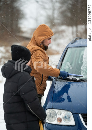Man clearing snow from car windshield with a child watching. Winter weather preparedness. 135996776